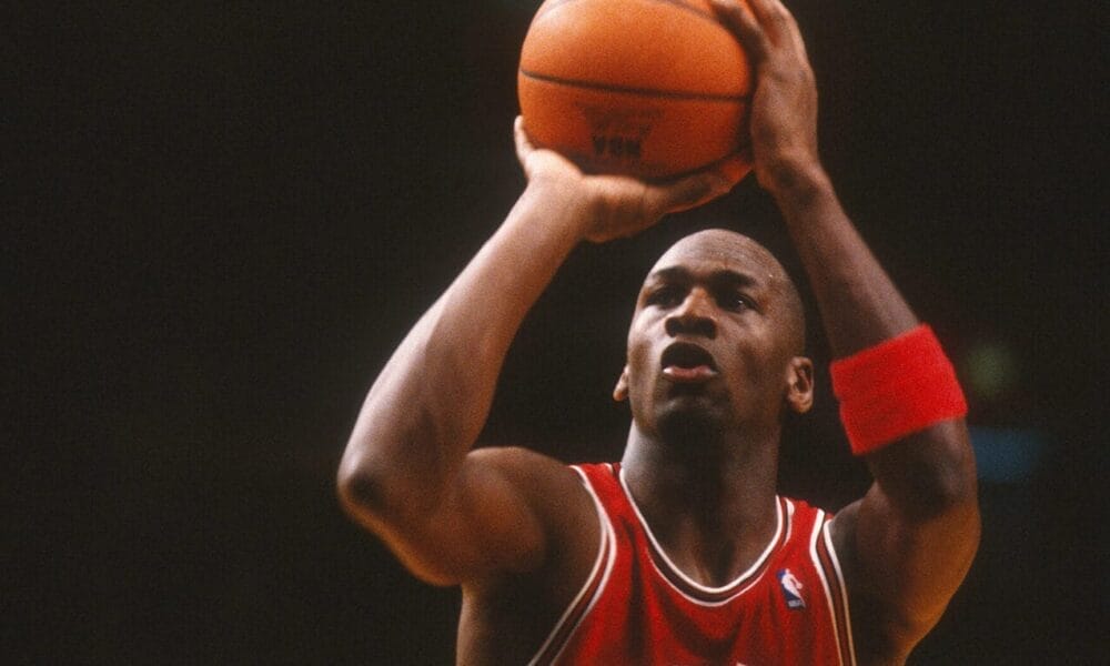 A basketball player in a red Bulls jersey, number 23, channels his inner Michael Jordan as he prepares to shoot a free throw during an NBA game. Clutching the ball with both hands and wearing a wristband on his left arm, he intensely focuses on the hoop against the dimly lit background.