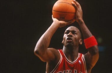 A basketball player in a red Bulls jersey, number 23, channels his inner Michael Jordan as he prepares to shoot a free throw during an NBA game. Clutching the ball with both hands and wearing a wristband on his left arm, he intensely focuses on the hoop against the dimly lit background.