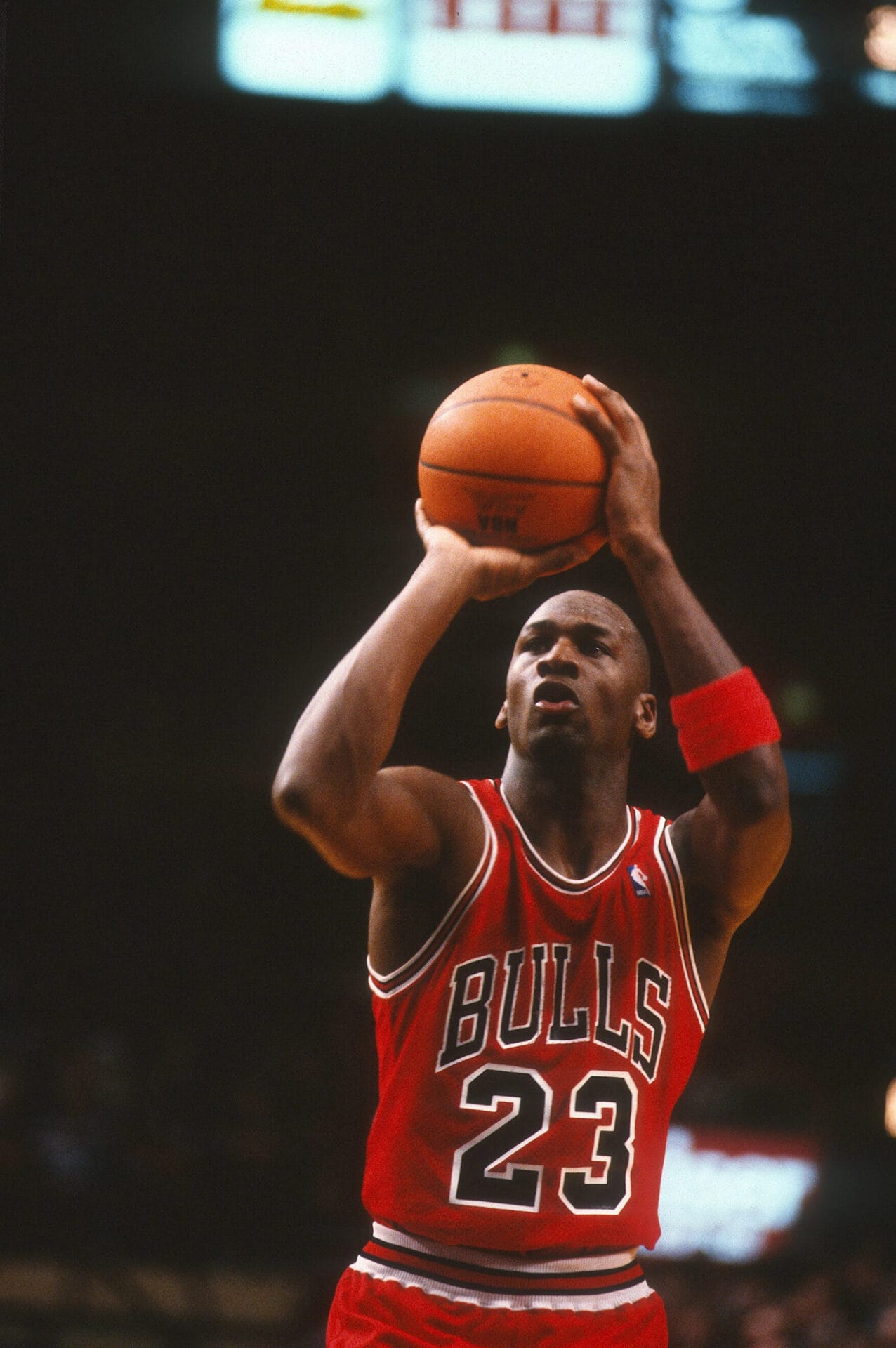 A basketball player in a red Bulls jersey, number 23, channels his inner Michael Jordan as he prepares to shoot a free throw during an NBA game. Clutching the ball with both hands and wearing a wristband on his left arm, he intensely focuses on the hoop against the dimly lit background.