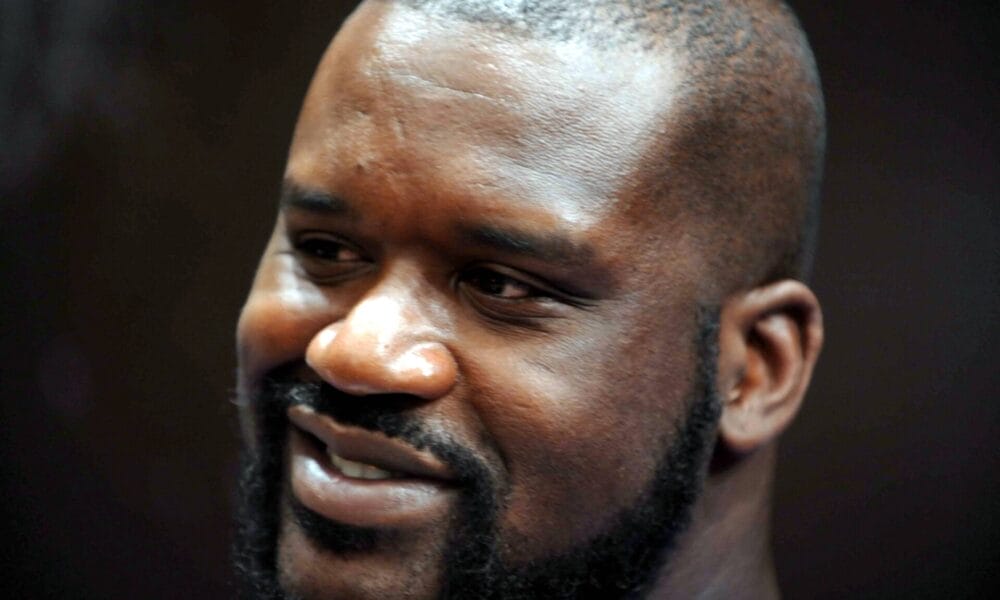 A person with a beard and a shaved head smiling, reminiscent of Shaquille O'Neal, captured in close-up against a dark background.