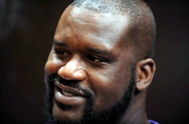 A person with a beard and a shaved head smiling, reminiscent of Shaquille O'Neal, captured in close-up against a dark background.