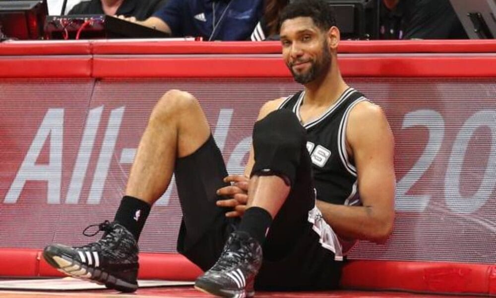 A basketball player in a black jersey, much like the iconic Tim Duncan during his illustrious NBA career, sits on the sideline of the court. Wearing black knee pads and shoes with white stripes, his hands rest on his knees as he gazes slightly to the side, framed by a backdrop of eager spectators.