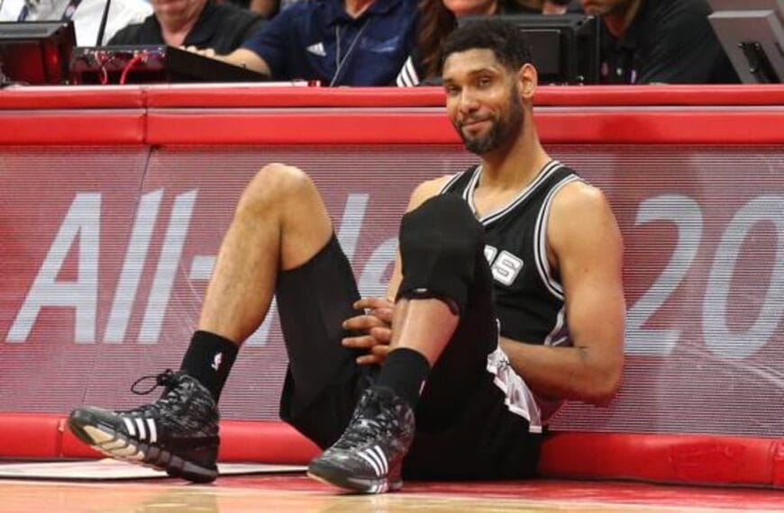 A basketball player in a black jersey, much like the iconic Tim Duncan during his illustrious NBA career, sits on the sideline of the court. Wearing black knee pads and shoes with white stripes, his hands rest on his knees as he gazes slightly to the side, framed by a backdrop of eager spectators.