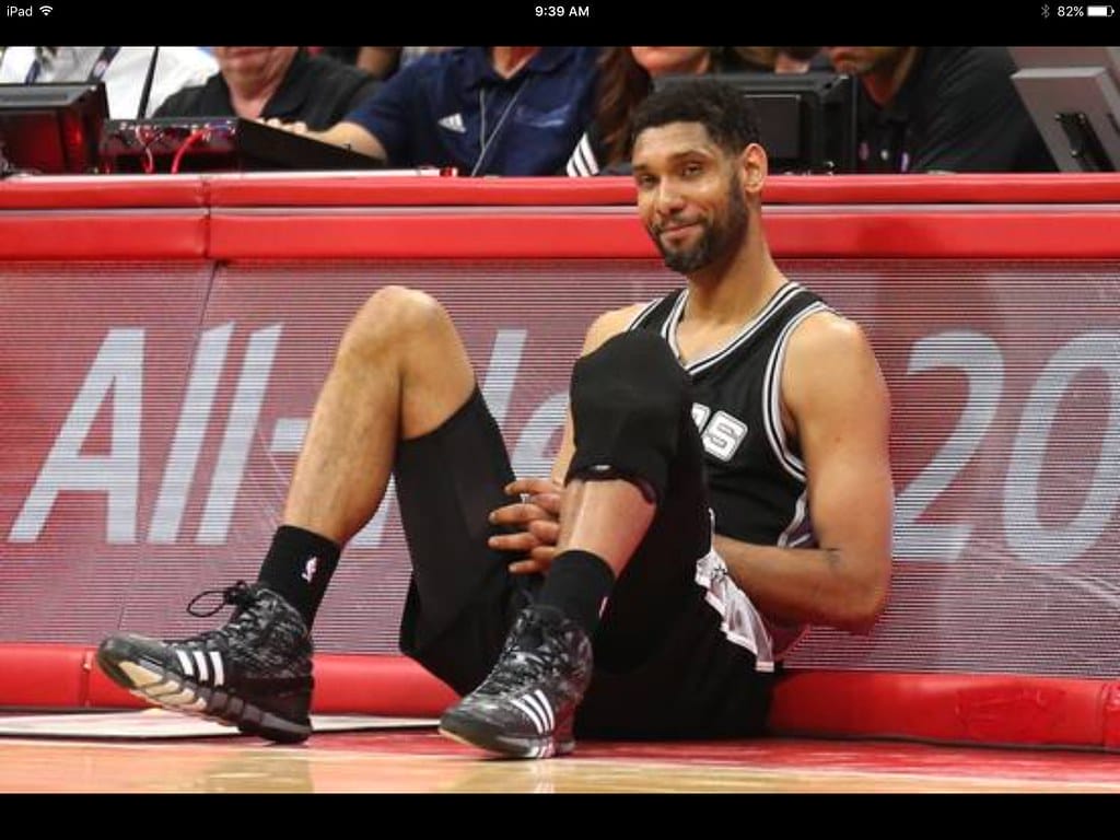 A basketball player in a black jersey, much like the iconic Tim Duncan during his illustrious NBA career, sits on the sideline of the court. Wearing black knee pads and shoes with white stripes, his hands rest on his knees as he gazes slightly to the side, framed by a backdrop of eager spectators.