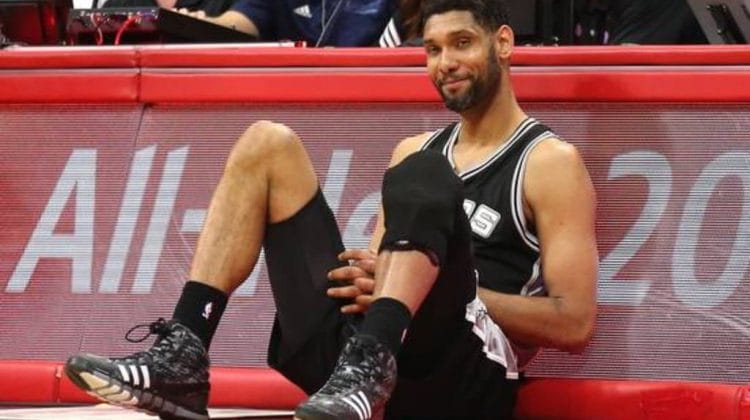 A basketball player in a black jersey, much like the iconic Tim Duncan during his illustrious NBA career, sits on the sideline of the court. Wearing black knee pads and shoes with white stripes, his hands rest on his knees as he gazes slightly to the side, framed by a backdrop of eager spectators.
