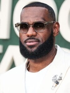 A person with a beard and sunglasses, reminiscent of LeBron James' iconic style, is wearing a white suit and a decorative pin. The background is blurred and green.