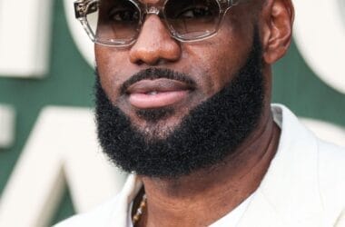 A person with a beard and sunglasses, reminiscent of LeBron James' iconic style, is wearing a white suit and a decorative pin. The background is blurred and green.