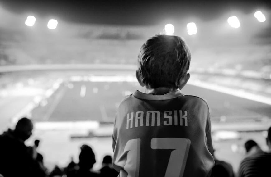 A young sports superfans with short hair, clad in a jersey emblazoned with "Hamsik" and number "17," sits enthralled in a stadium. The black-and-white scene is bathed in the glow of stadium lights as they watch the event unfold.
