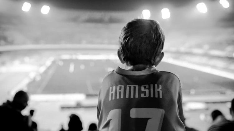 A young sports superfans with short hair, clad in a jersey emblazoned with "Hamsik" and number "17," sits enthralled in a stadium. The black-and-white scene is bathed in the glow of stadium lights as they watch the event unfold.