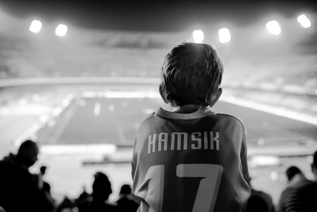 A young sports superfans with short hair, clad in a jersey emblazoned with "Hamsik" and number "17," sits enthralled in a stadium. The black-and-white scene is bathed in the glow of stadium lights as they watch the event unfold.
