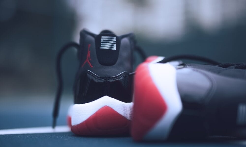 A close-up of a pair of sneakers on a surface evokes the essence of 90s basketball. The shoes, primarily black with red and white accents, display a logo on the heel. While one stands upright, the other lies on its side against a blurred background.