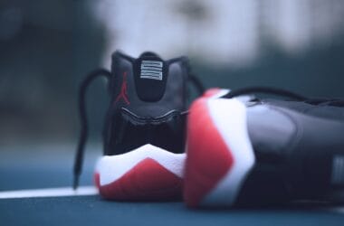 A close-up of a pair of sneakers on a surface evokes the essence of 90s basketball. The shoes, primarily black with red and white accents, display a logo on the heel. While one stands upright, the other lies on its side against a blurred background.