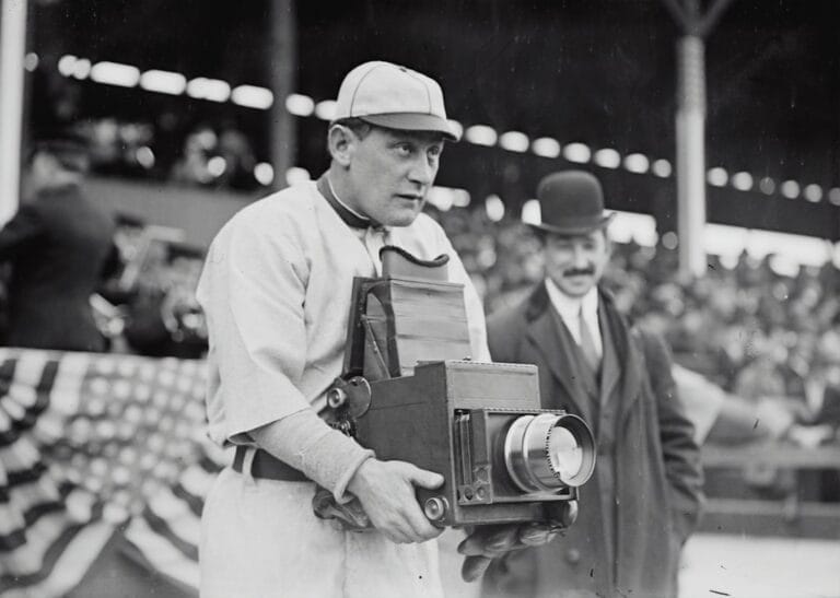 A man in a vintage baseball uniform holds a large box camera at the bustling stadium, capturing the essence of baseball legends. Another man, donning a suit and bowler hat, stands behind him. The scene is framed by the crowd's excitement and an American flag bunting waving proudly.