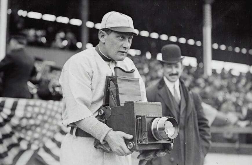 A man in a vintage baseball uniform holds a large box camera at the bustling stadium, capturing the essence of baseball legends. Another man, donning a suit and bowler hat, stands behind him. The scene is framed by the crowd's excitement and an American flag bunting waving proudly.