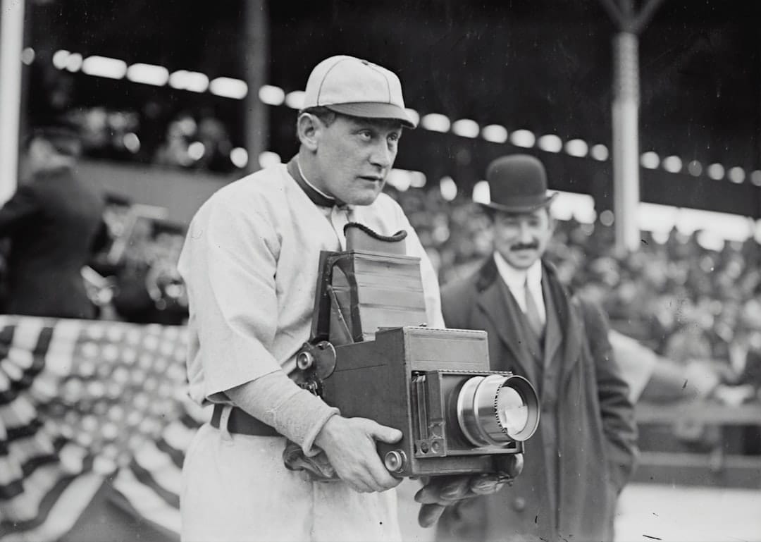 A man in a vintage baseball uniform holds a large box camera at the bustling stadium, capturing the essence of baseball legends. Another man, donning a suit and bowler hat, stands behind him. The scene is framed by the crowd's excitement and an American flag bunting waving proudly.
