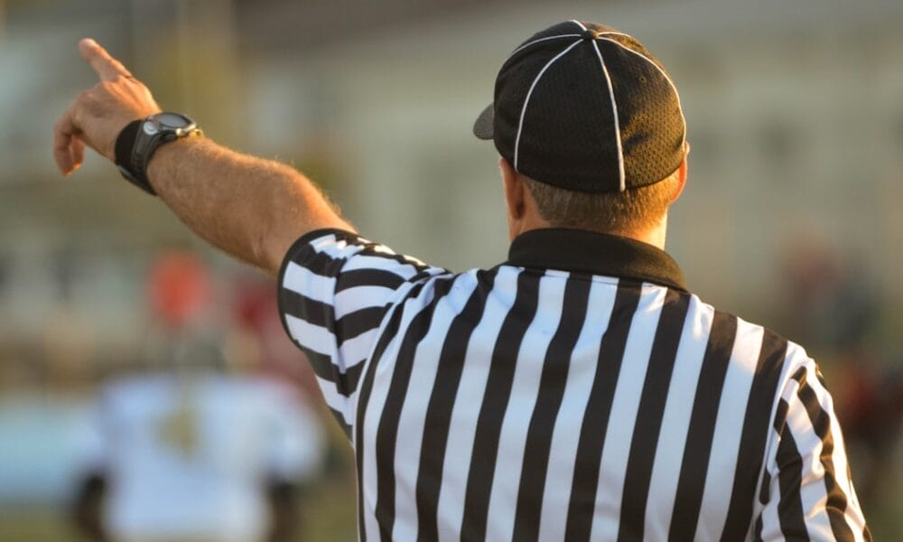 A referee, adhering to NFL regulations, is seen from behind in a black-and-white striped shirt and black cap, raising one arm and pointing. The blurred background reveals a sports field bustling with players.