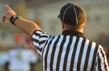 A referee, adhering to NFL regulations, is seen from behind in a black-and-white striped shirt and black cap, raising one arm and pointing. The blurred background reveals a sports field bustling with players.