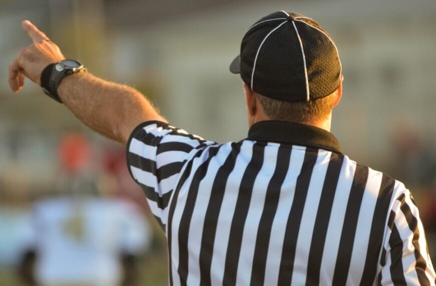 A referee, adhering to NFL regulations, is seen from behind in a black-and-white striped shirt and black cap, raising one arm and pointing. The blurred background reveals a sports field bustling with players.