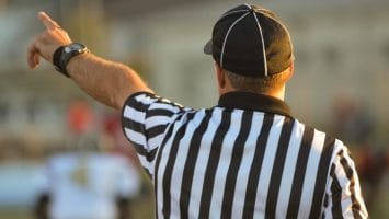 A referee, adhering to NFL regulations, is seen from behind in a black-and-white striped shirt and black cap, raising one arm and pointing. The blurred background reveals a sports field bustling with players.
