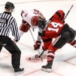 A referee prepares to drop the puck as two ice hockey players, one in a white jersey and the other in a red and black jersey, face off. Poised with their sticks near the ice, they embody the unwritten rules in the NHL, ready to start the play.