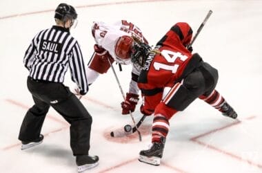 A referee prepares to drop the puck as two ice hockey players, one in a white jersey and the other in a red and black jersey, face off. Poised with their sticks near the ice, they embody the unwritten rules in the NHL, ready to start the play.