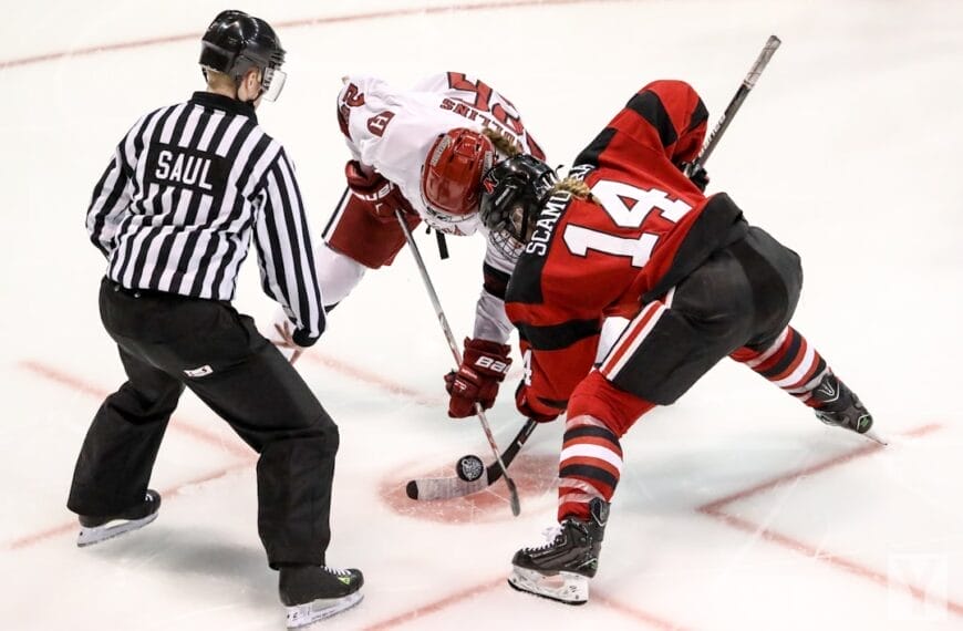 A referee prepares to drop the puck as two ice hockey players, one in a white jersey and the other in a red and black jersey, face off. Poised with their sticks near the ice, they embody the unwritten rules in the NHL, ready to start the play.