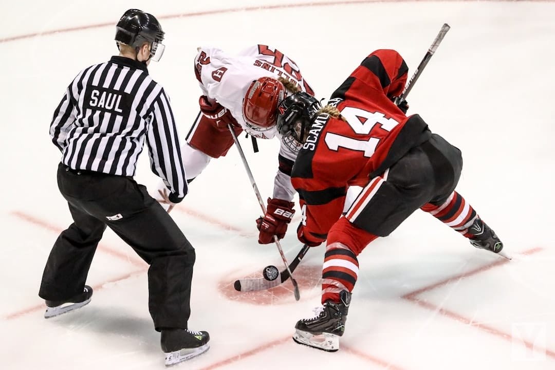 A referee prepares to drop the puck as two ice hockey players, one in a white jersey and the other in a red and black jersey, face off. Poised with their sticks near the ice, they embody the unwritten rules in the NHL, ready to start the play.