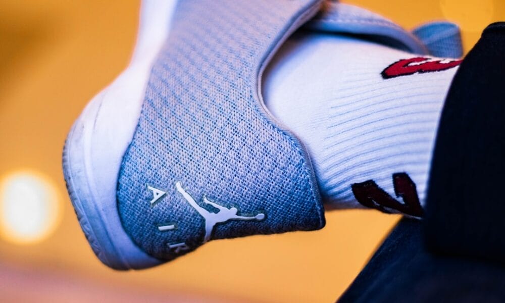 Close-up of a foot in a gray sneaker with a white logo and the word "AIR," reminiscent of 90s basketball impact. The person sports a white sock adorned with red and black designs, set against a softly blurred background with warm lighting.