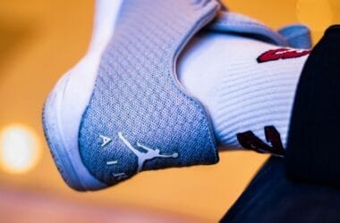 Close-up of a foot in a gray sneaker with a white logo and the word "AIR," reminiscent of 90s basketball impact. The person sports a white sock adorned with red and black designs, set against a softly blurred background with warm lighting.