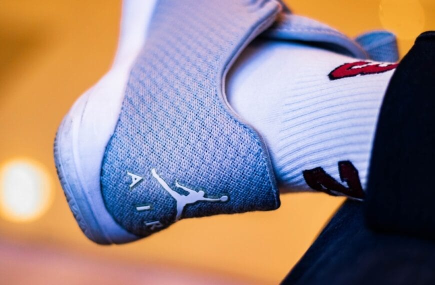 Close-up of a foot in a gray sneaker with a white logo and the word "AIR," reminiscent of 90s basketball impact. The person sports a white sock adorned with red and black designs, set against a softly blurred background with warm lighting.