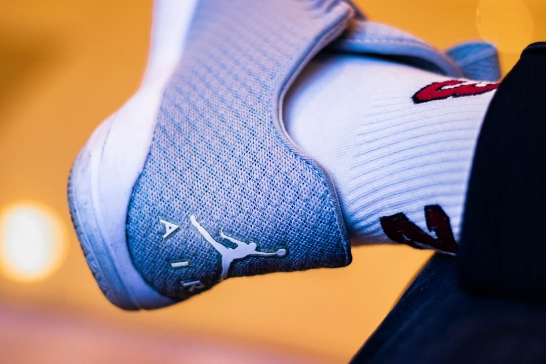 Close-up of a foot in a gray sneaker with a white logo and the word "AIR," reminiscent of 90s basketball impact. The person sports a white sock adorned with red and black designs, set against a softly blurred background with warm lighting.