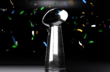 A silver trophy resembling the Vince Lombardi Trophy is centered against a dark background, celebrating the greatest NFL players of all time. Colorful confetti—blue, yellow, green, and white—floats through the air around this iconic symbol of victory.