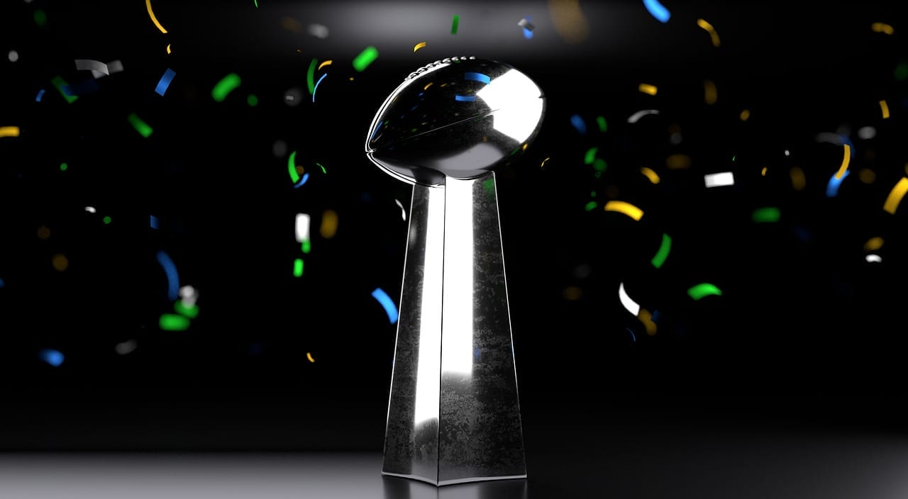 A silver trophy resembling the Vince Lombardi Trophy is centered against a dark background, celebrating the greatest NFL players of all time. Colorful confetti—blue, yellow, green, and white—floats through the air around this iconic symbol of victory.