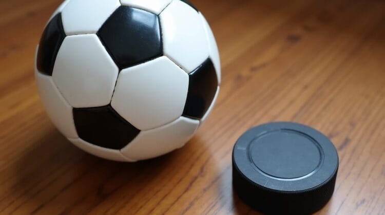 A black and white soccer ball and a black hockey puck rest side by side on a wooden surface, subtly highlighting the MLS vs NHL viewership rivalry. Soft light shines on them, emphasizing their distinct shapes and textures.