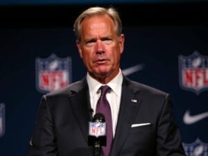 A middle-aged man in a dark suit and purple tie stands at a podium with an NFL Analytics microphone, speaking against a backdrop of repeating NFL logos. He has light hair, a serious expression, and wears a white pocket square.