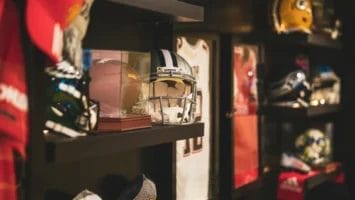 A wall display features shelves with American football helmets, sports memorabilia, photographs of famous gridiron players, and framed jerseys. The scene is warmly lit, highlighting helmets and collectibles arranged neatly on the dark shelves.