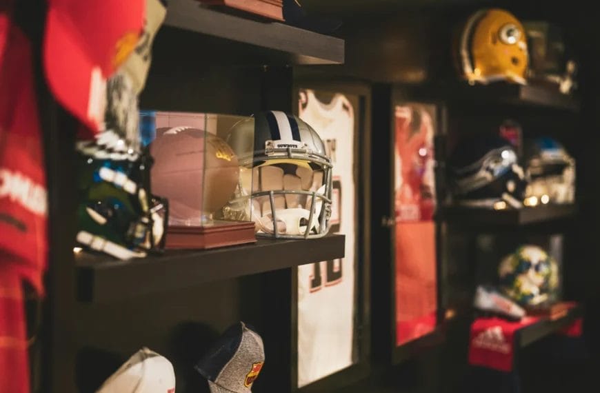 A wall display features shelves with American football helmets, sports memorabilia, photographs of famous gridiron players, and framed jerseys. The scene is warmly lit, highlighting helmets and collectibles arranged neatly on the dark shelves.
