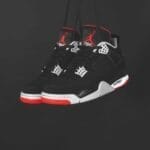 A pair of black, white, and red Air Jordan sneakers hang by their laces against a dark background. Featuring the iconic Jumpman Logo, white lace holders, red accents, and a visible air unit in the midsole.