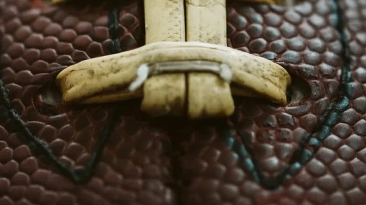 Close-up of a brown American football, focusing on the textured, pebbled leather surface and tan, slightly worn laces—crafted to comply with national football league rules—with visible stitching running vertically through the center of the frame.