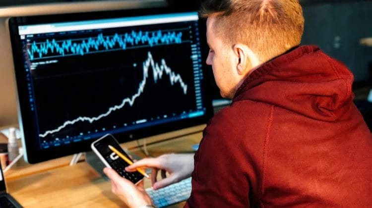 A man in a maroon hoodie sits at a desk, analyzing nba basketball analytics with sharp rises and falls on a large computer monitor. He holds a smartphone and pencil, focusing intently on the data. A keyboard and notebook sit on the desk.