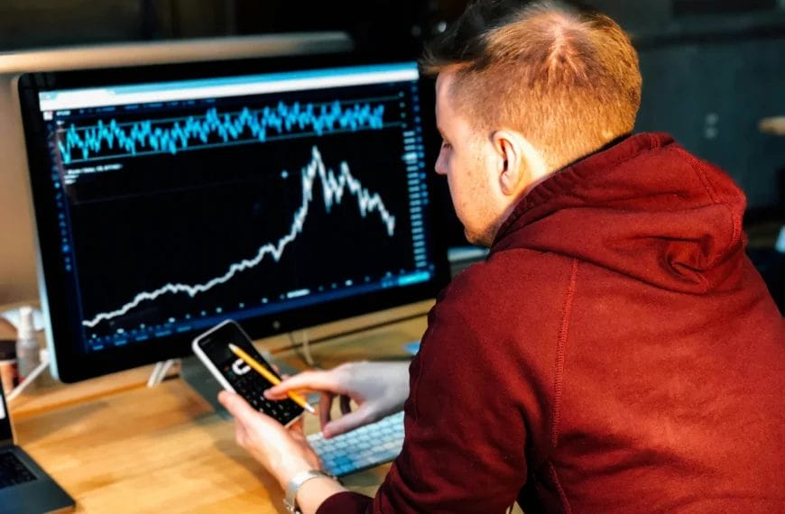 A man in a maroon hoodie sits at a desk, analyzing nba basketball analytics with sharp rises and falls on a large computer monitor. He holds a smartphone and pencil, focusing intently on the data. A keyboard and notebook sit on the desk.