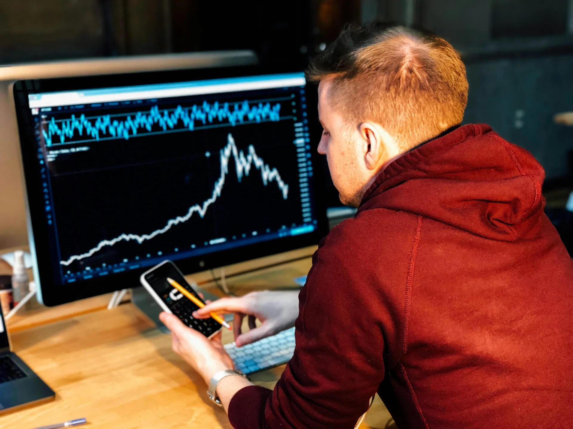 A man in a maroon hoodie sits at a desk, analyzing nba basketball analytics with sharp rises and falls on a large computer monitor. He holds a smartphone and pencil, focusing intently on the data. A keyboard and notebook sit on the desk.
