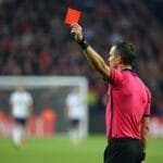 A soccer referee in a pink shirt holds up a red card on the field, signaling a player’s expulsion—a scene often linked to controversial sports moments. The blurred background shows players in white uniforms and a crowd in the stands.