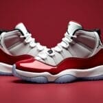 A pair of high-top sneakers with glossy red mudguards, white mesh uppers, black accents, and translucent icy blue soles pays tribute to Michael Jordan—a cultural icon—displayed against a solid red background with the iconic Jumpman logo on the ankle.