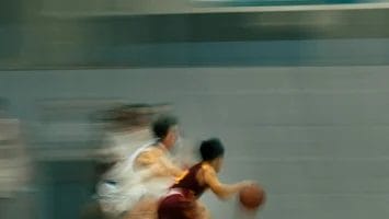 A blurred action shot of a youth basketball game shows a player in a maroon uniform, known for his impressive player efficiency rating, dribbling the ball quickly past defenders in white on an indoor court, emphasizing speed and movement.