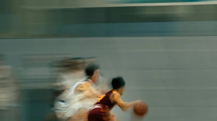 A blurred action shot of a youth basketball game shows a player in a maroon uniform, known for his impressive player efficiency rating, dribbling the ball quickly past defenders in white on an indoor court, emphasizing speed and movement.