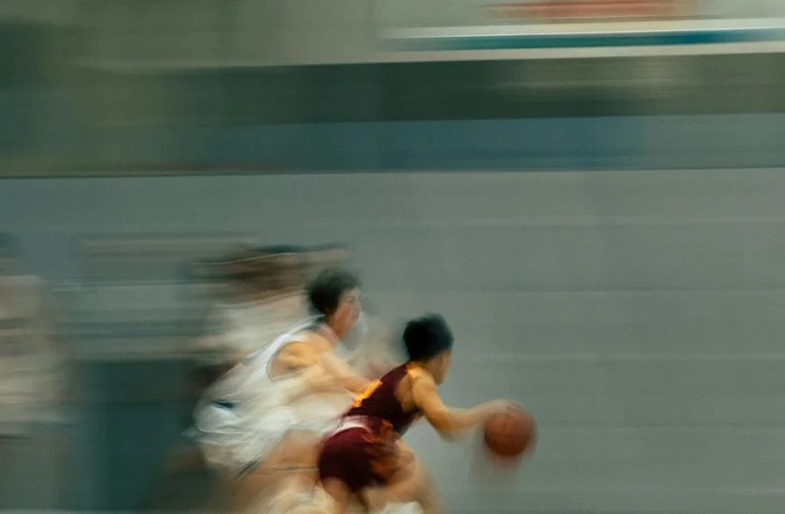 A blurred action shot of a youth basketball game shows a player in a maroon uniform, known for his impressive player efficiency rating, dribbling the ball quickly past defenders in white on an indoor court, emphasizing speed and movement.