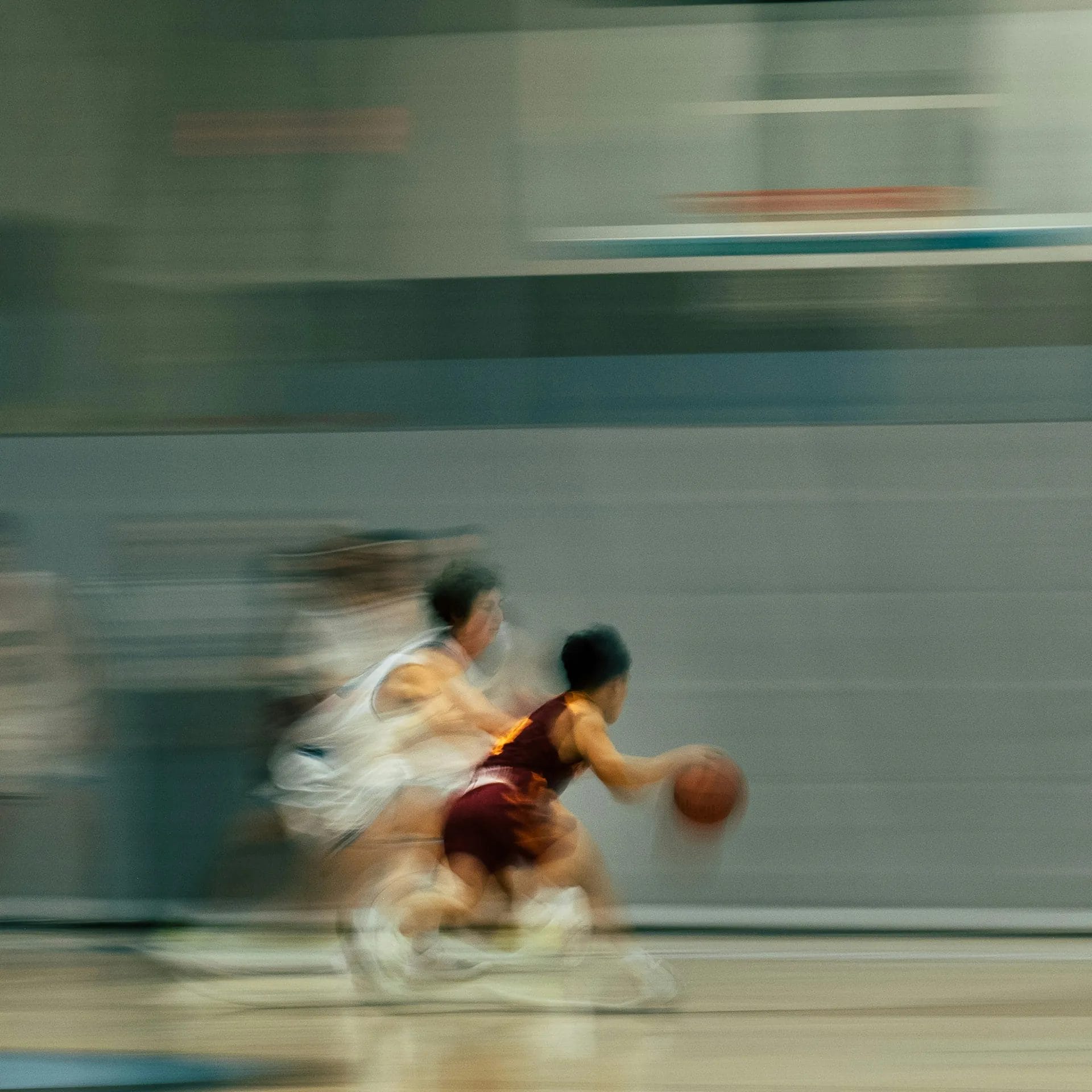 A blurred action shot of a youth basketball game shows a player in a maroon uniform, known for his impressive player efficiency rating, dribbling the ball quickly past defenders in white on an indoor court, emphasizing speed and movement.