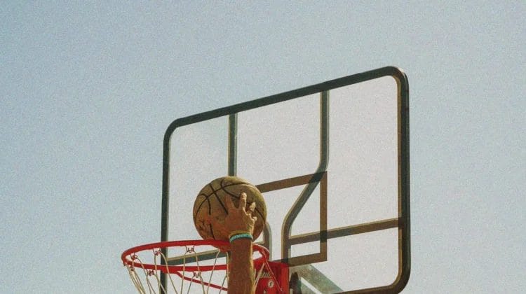 A shirtless person in a backward cap jumps to dunk a basketball into an outdoor hoop against a bright, clear blue sky. The image is shot from below, capturing the action and the ball just above the rim.