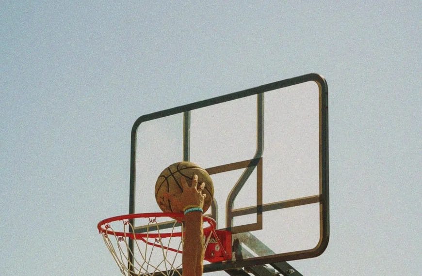 A shirtless person in a backward cap jumps to dunk a basketball into an outdoor hoop against a bright, clear blue sky. The image is shot from below, capturing the action and the ball just above the rim.